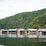 Na imagem há o Rio Amazonas, com mata aberta ao fundo e casas bem simples que estão fincadas dentro do rio.